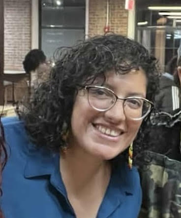 An image of a person smiling with dark curly hair, blue shirt and glasses.